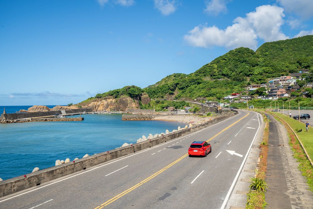 A red car driving along a coastal road by the sea